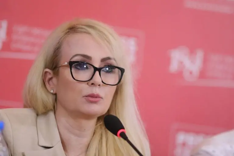 Eine Frau mit blonden Haaren und Brille spricht bei einer Konferenz, mit einem Mikrofon vor einem roten Hintergrund.