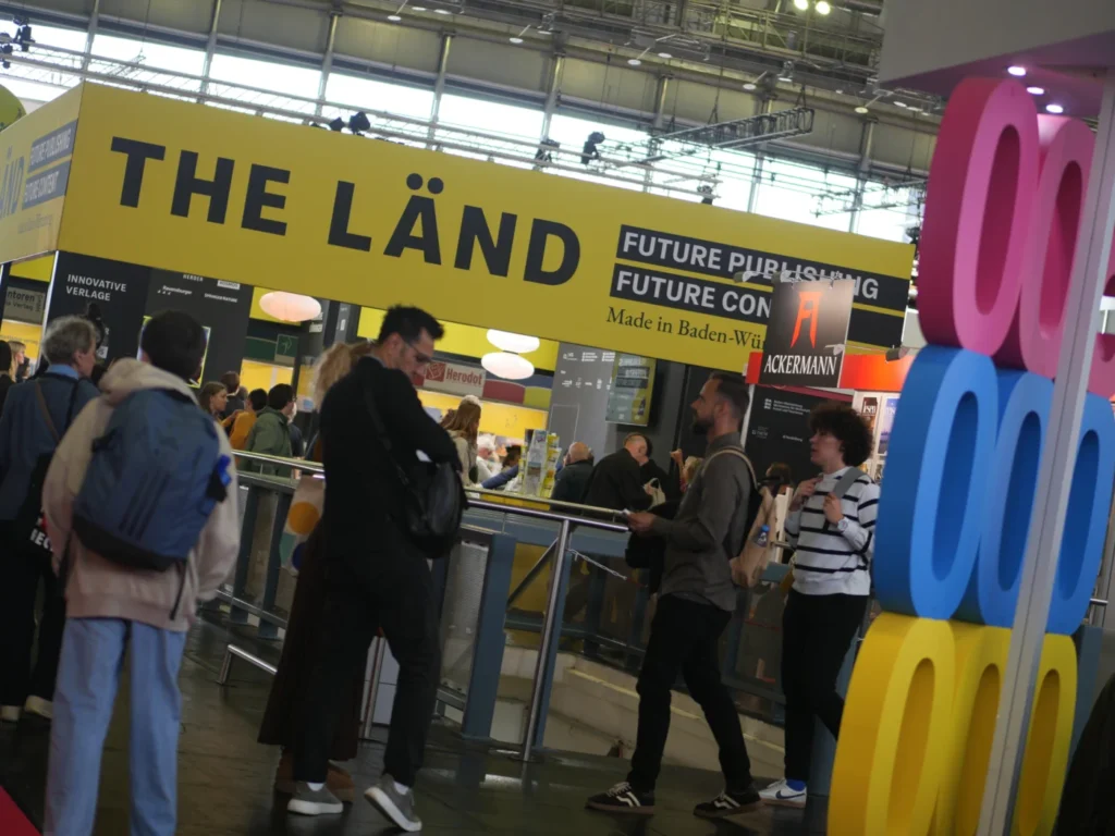 Menschenmenge am Stand 'THE LÄND' auf der Buchmesse mit bunten Dekorationen und Informationsständen im Hintergrund.