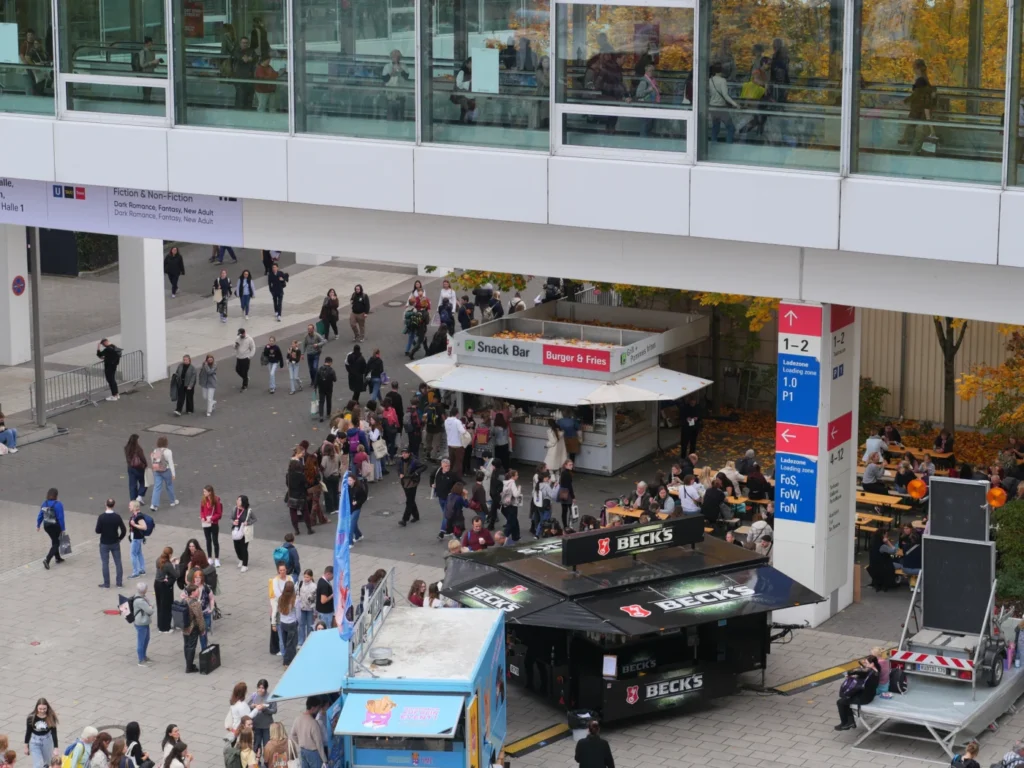 Eine belebte Aufnahme der Außenbereiche der Buchmesse in Frankfurt, mit Ständen, Besuchern und einem Essensstand, umgeben von herbstlichen Bäumen.