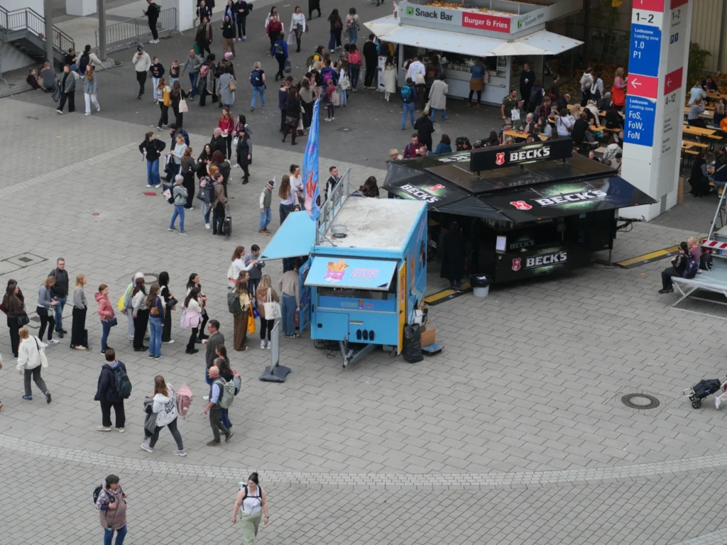 Blick auf eine belebte Veranstaltungsfläche mit mehreren Menschen, die sich nahe einem blauen Verkaufsstand und einem schwarzen Getränkeverkaufsstand aufhalten. Im Hintergrund sind weitere Stände und Tische sichtbar.