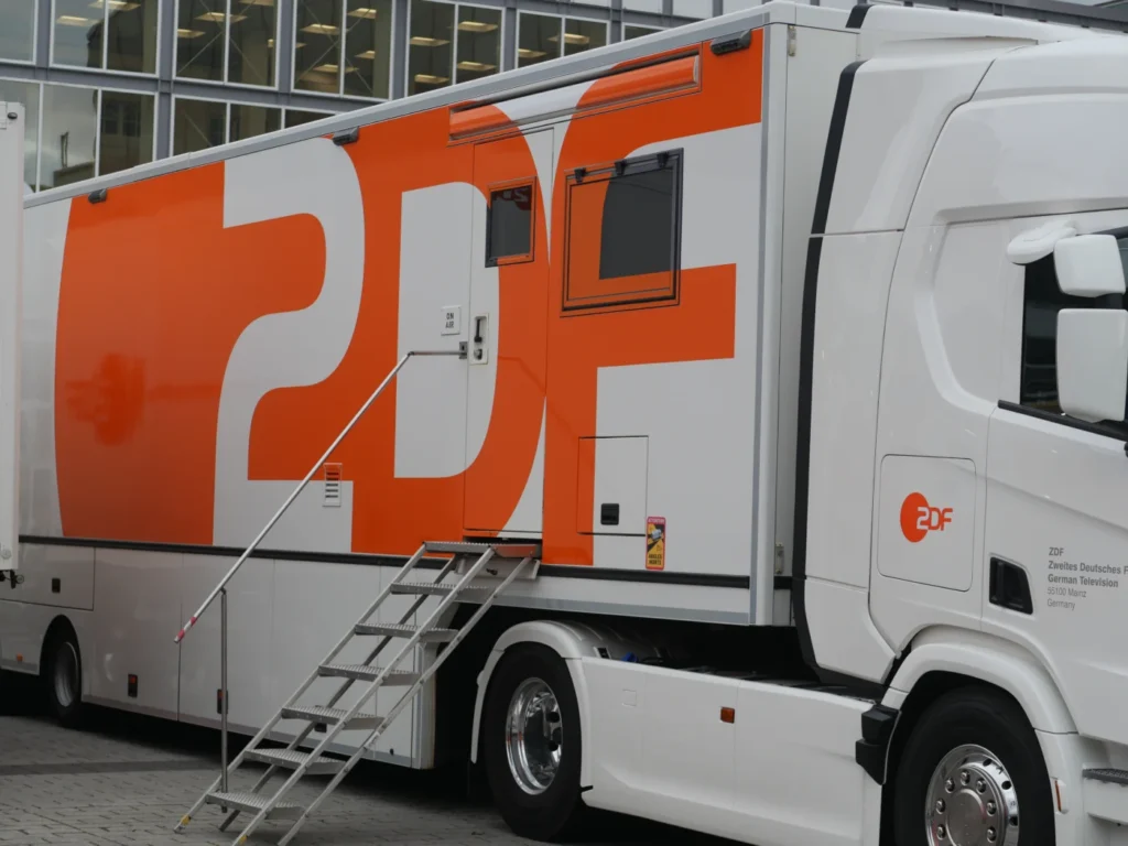 Ein Fahrzeug der deutschen Rundfunkanstalt ZDF mit großem orangefarbenen Logo, ausgestattet mit Treppen und Sichtfenster.