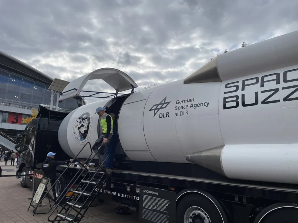 Ein Lastwagen mit einem Modell einer Raumfähre, das von der Deutschen Raumfahrtagentur DLR präsentiert wird. Eine Person geht eine Treppe hinauf zum Eingang der Fähre, während eine andere Person auf dem Boden steht und auf die Fähre zeigt. Der Himmel ist bewölkt.