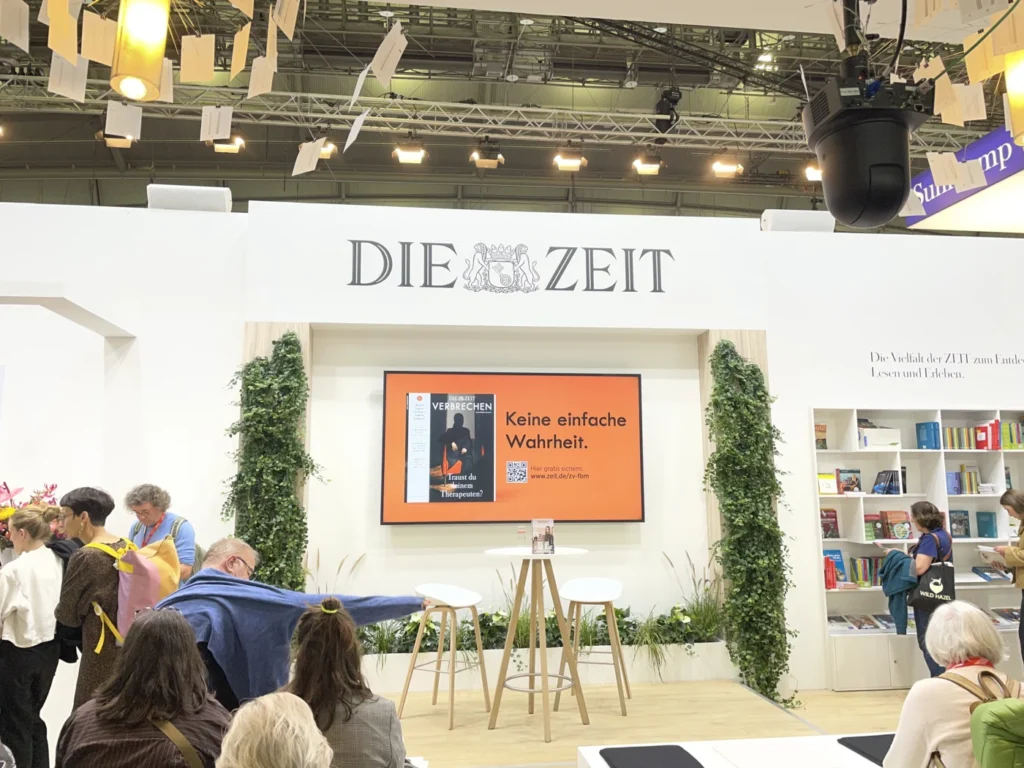 Messestand von DIE ZEIT auf der Buchmesse, mit einem Bildschirm, der das Buch "Verbrechen" bewirbt. Besucher stehen in der Nähe, um sich zu informieren. Die Dekoration umfasst Pflanzen und Bücherregale im Hintergrund.