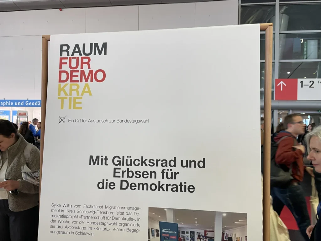 Ein Hinweisschild mit dem Text 'RAUM FÜR DEMOKRATIE' zur Bundestagswahl, umgeben von Besuchern auf der Buchmesse in Frankfurt.